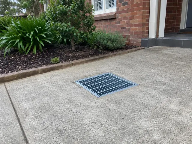 Concrete driveway with a seated stormwater grate beside a brick house and garden bed in Thornleigh